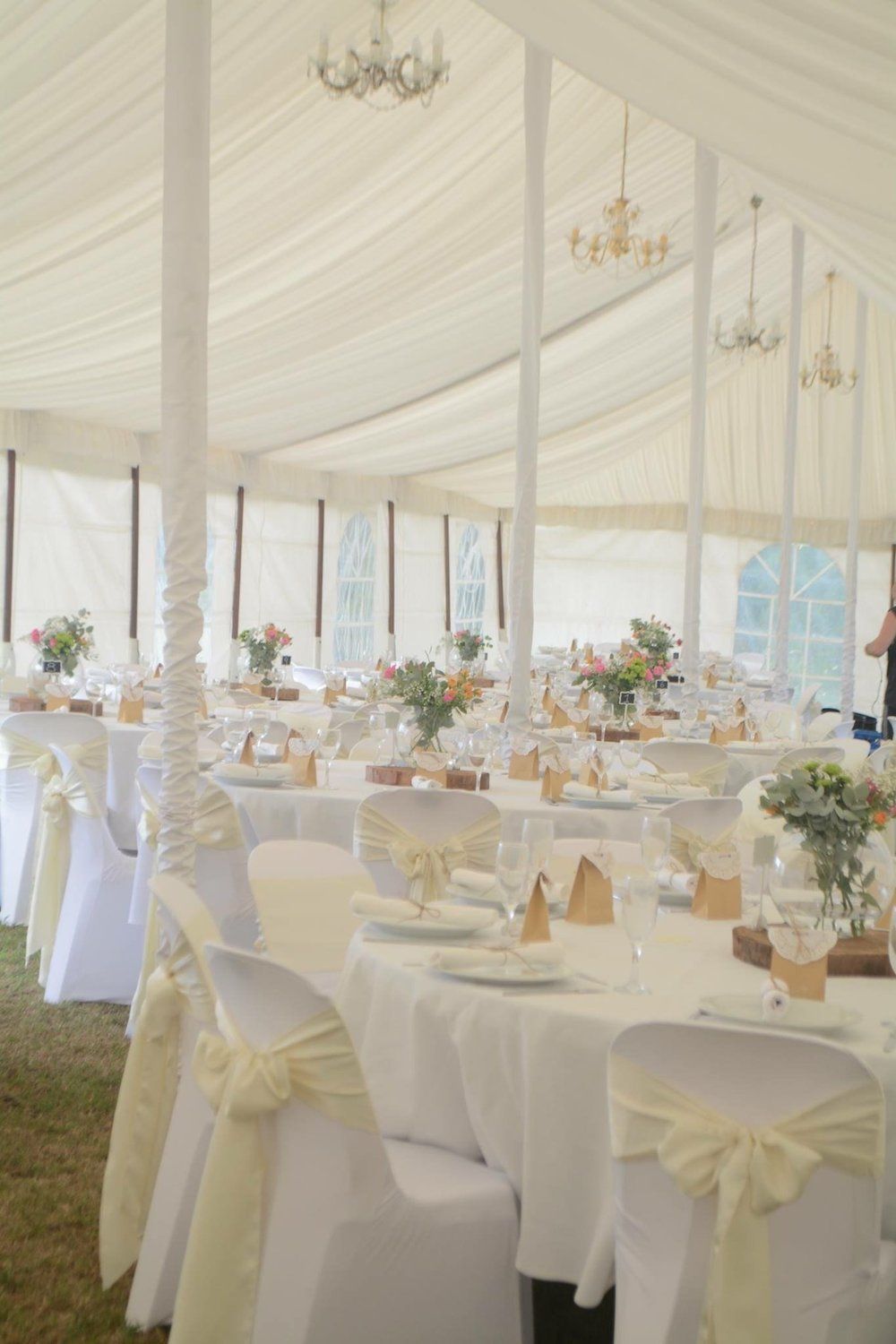 Wedding reception tent with round tables, white linens, floral centerpieces, and chandeliers.