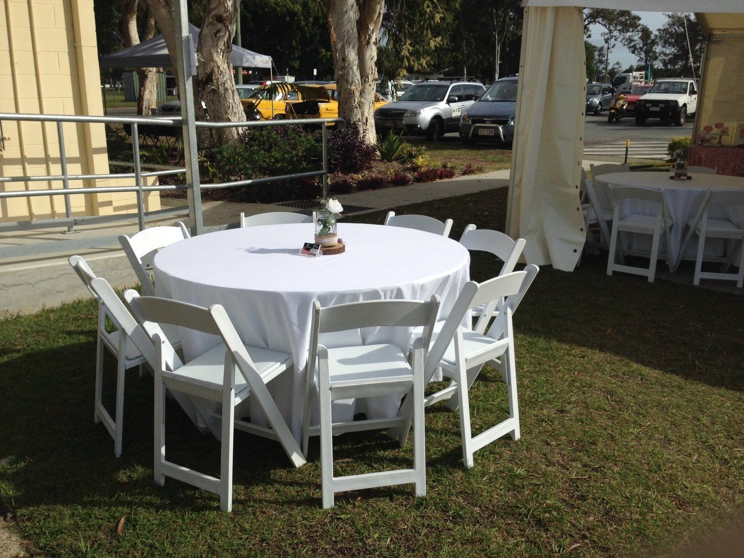 A round table with a white tablecloth and white folding chairs is set up on grass outdoors.