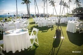 Outdoor wedding reception with white-covered tables on green grass; tiki torches, ocean view, palm trees in the background.