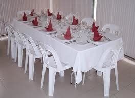 A white tablecloth set with plates, silverware, napkins, and glasses on a long table with white plastic chairs.