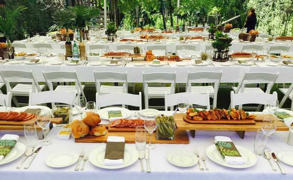 Long tables set for a meal outdoors, white chairs, various food items, green foliage background.