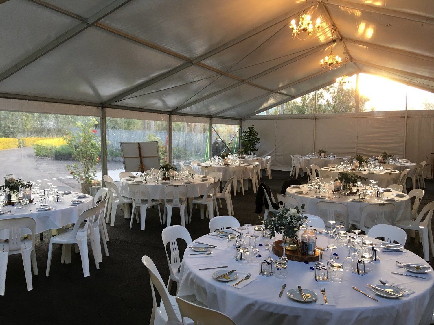 Inside a white tent, round tables with place settings are arranged for a catered event, with sunlight visible.