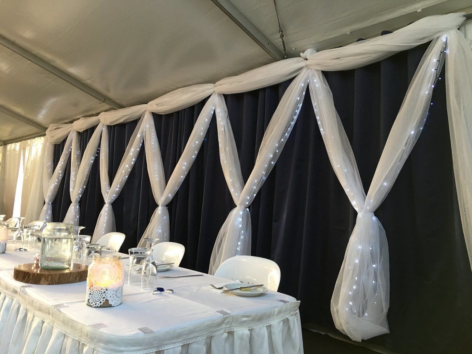 Blue-draped wall decorated with white fabric swags and string lights in a tent. Table set for a gathering.