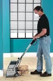 Man sanding hardwood floor, wearing mask, in room with teal walls.