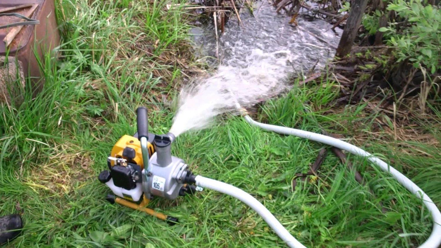 A water pump spraying water onto grass. The pump is yellow and silver with a white hose.
