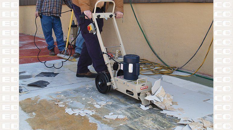 Man using floor scraper to remove flooring on a patio, another person nearby; outdoors.