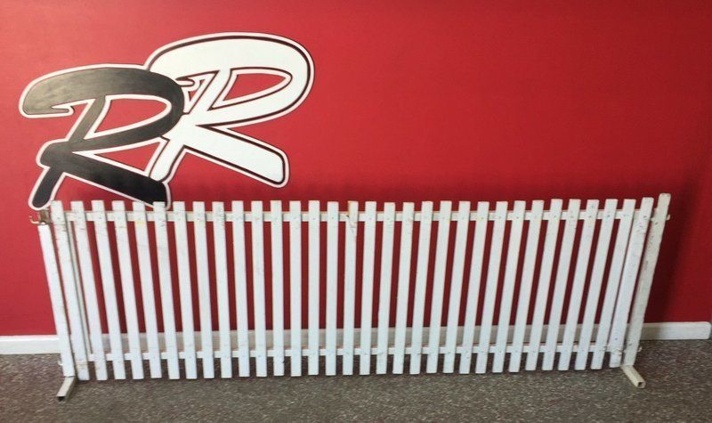White picket fence leaning against a red wall with a black and white logo.
