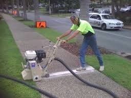 Man in yellow shirt using a sidewalk cleaning machine on a paved path beside a road.