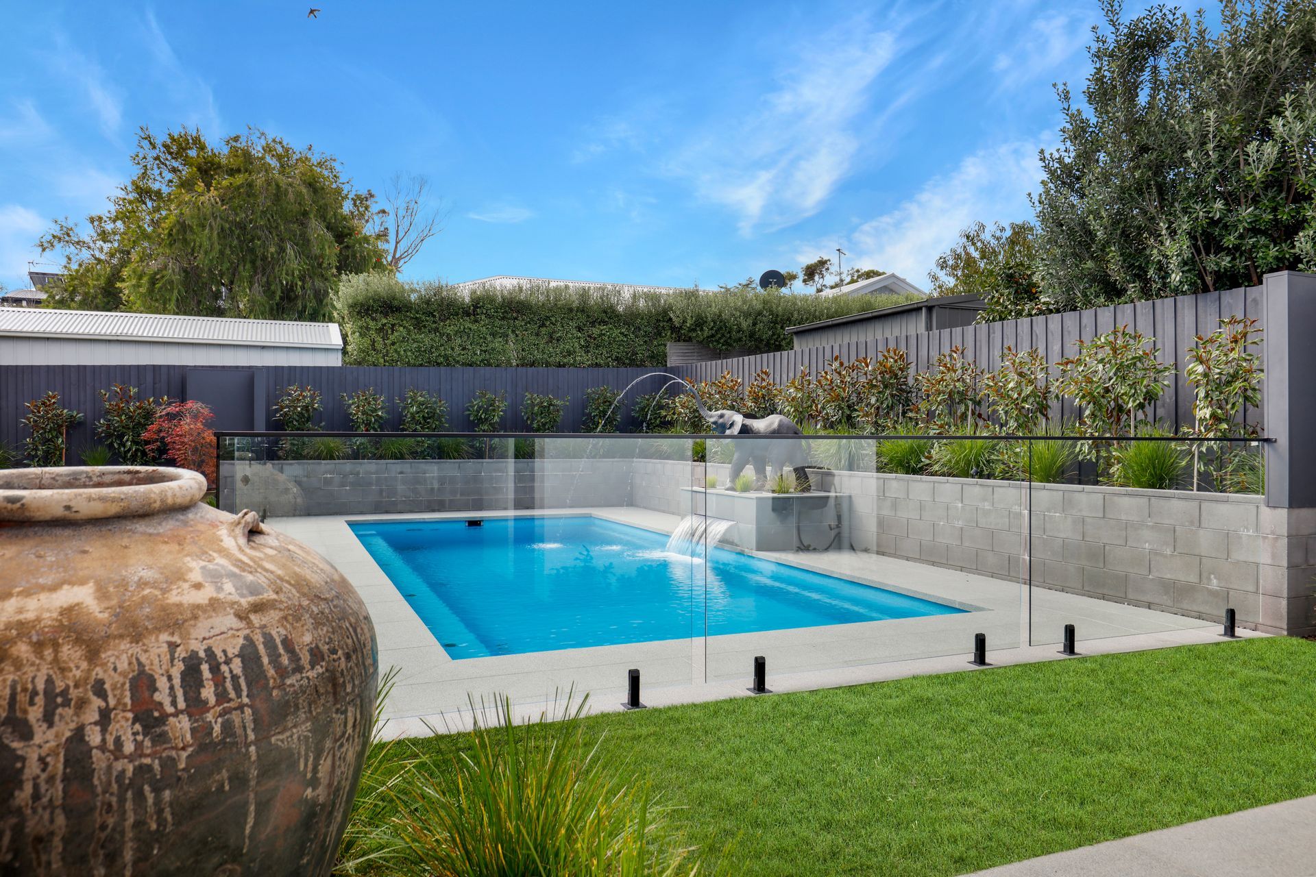 Backyard pool with glass fencing, green lawn, and a large clay pot under a blue sky