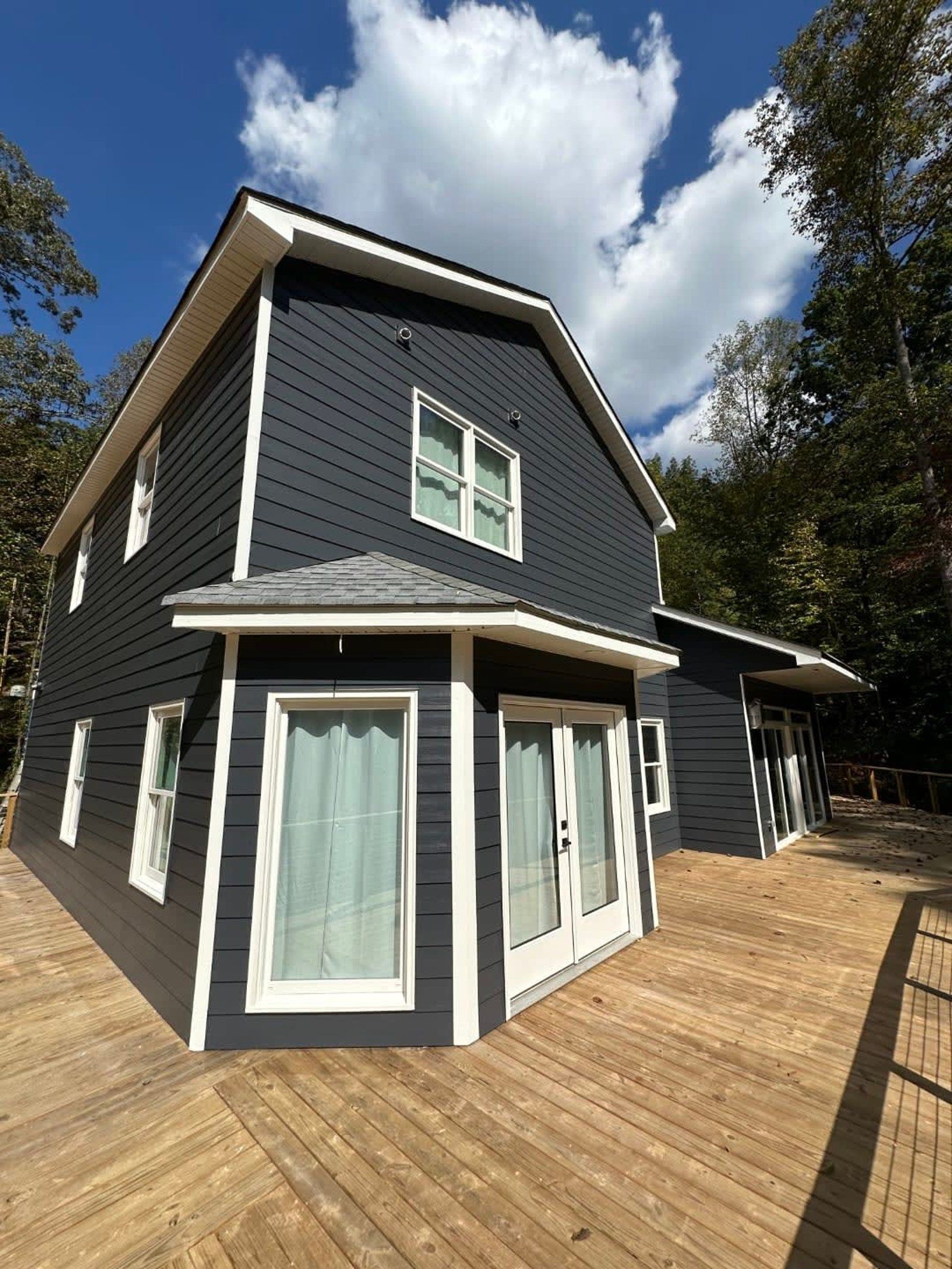 Two-story dark blue house with white trim, deck, and windows. Blue sky and trees in the background.