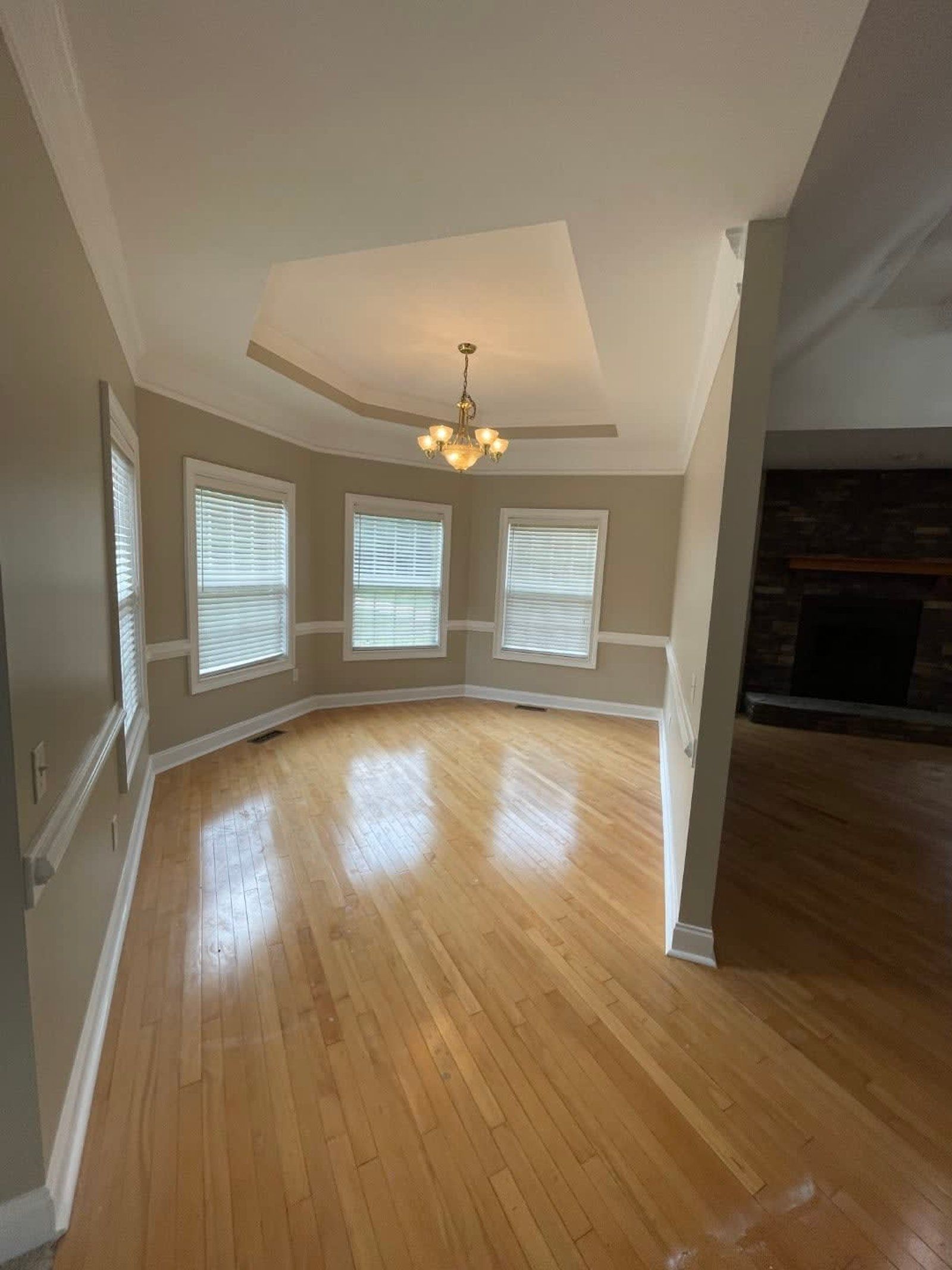 Wooden-floored room with tan walls, arched windows, and a raised ceiling with a chandelier. Fireplace visible in the adjacent room.
