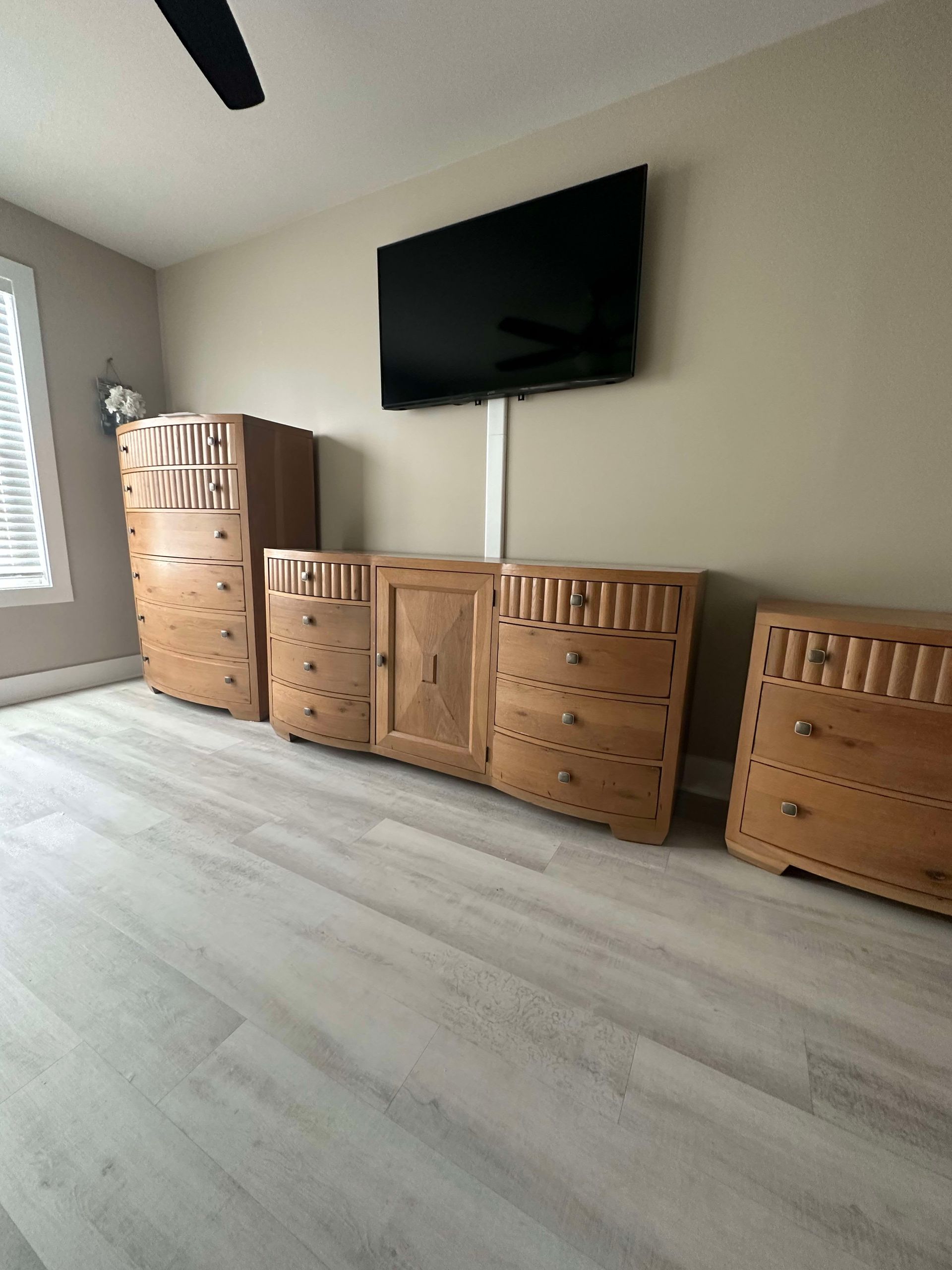 Bedroom furniture set: tall dresser, TV, wide cabinet, nightstand. Light wood, beige wall, light wood floor.