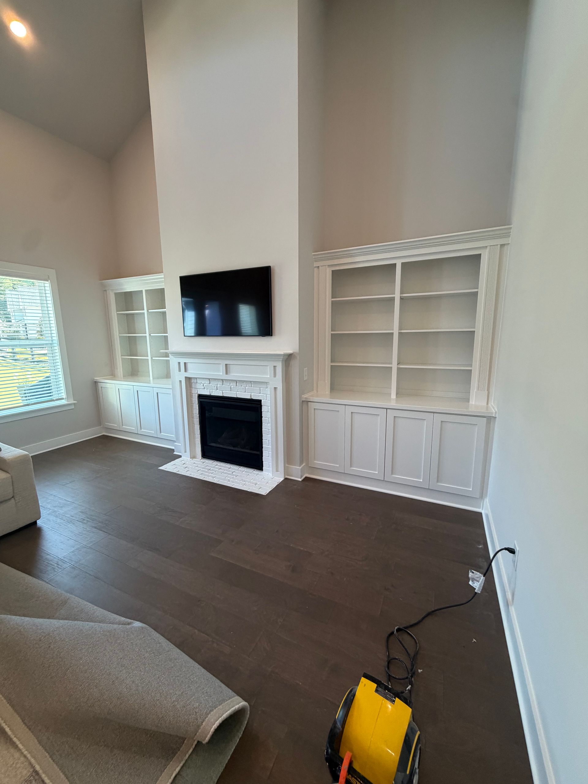 Bright living room with white built-in shelves, fireplace, TV, dark wood floor, and a yellow item in foreground.