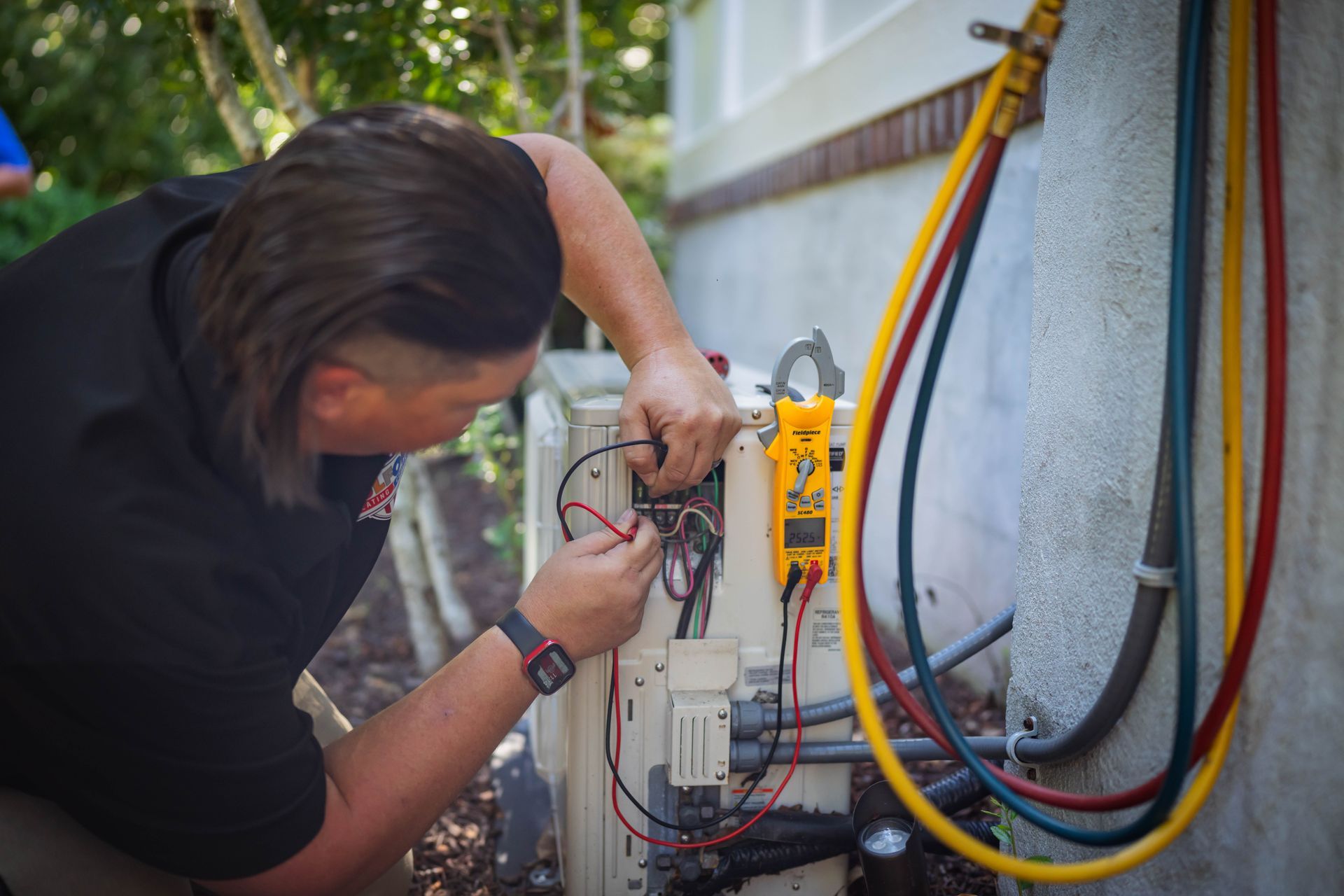 HVAC technician using multimeter on outdoor unit, connecting wires. Yellow, blue, and red hoses are present.