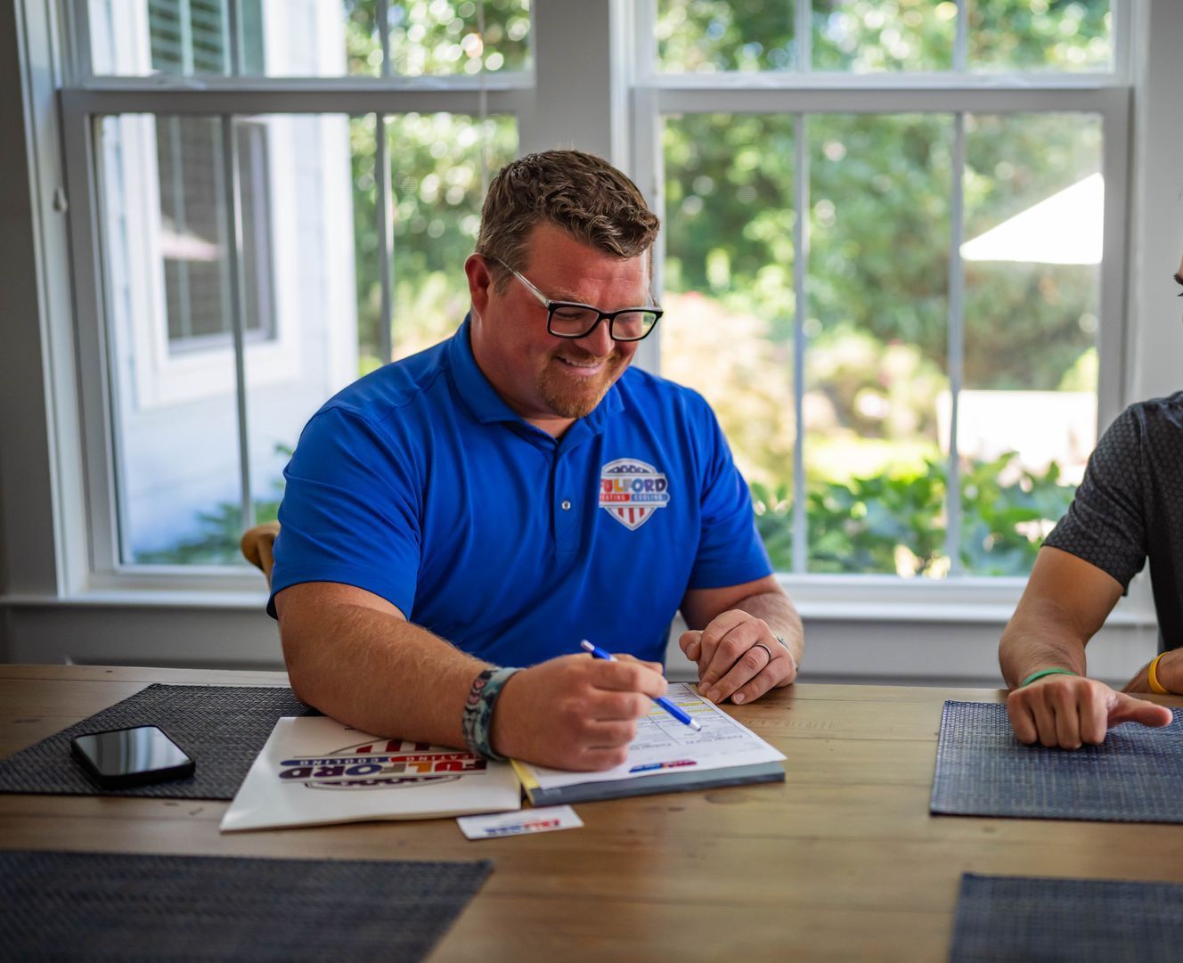 Man in blue shirt, glasses, signing paperwork at a table with another person, near a window.