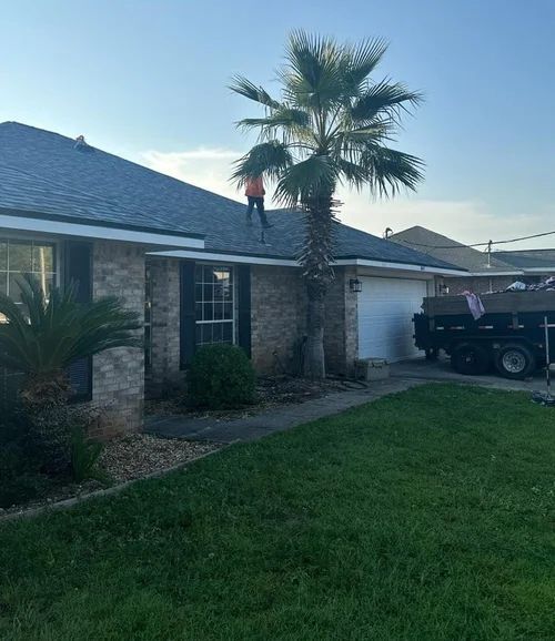 A person in an orange shirt stands on a residential roof, trimming a palm tree near a garage and a dump trailer.