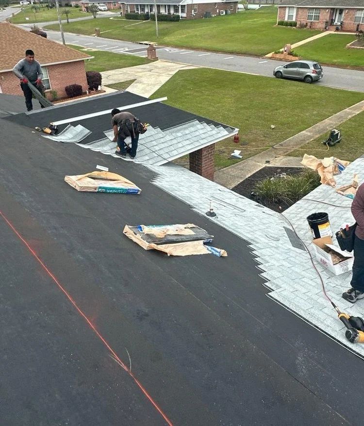 Three workers install shingles on a residential roof in a neighborhood.