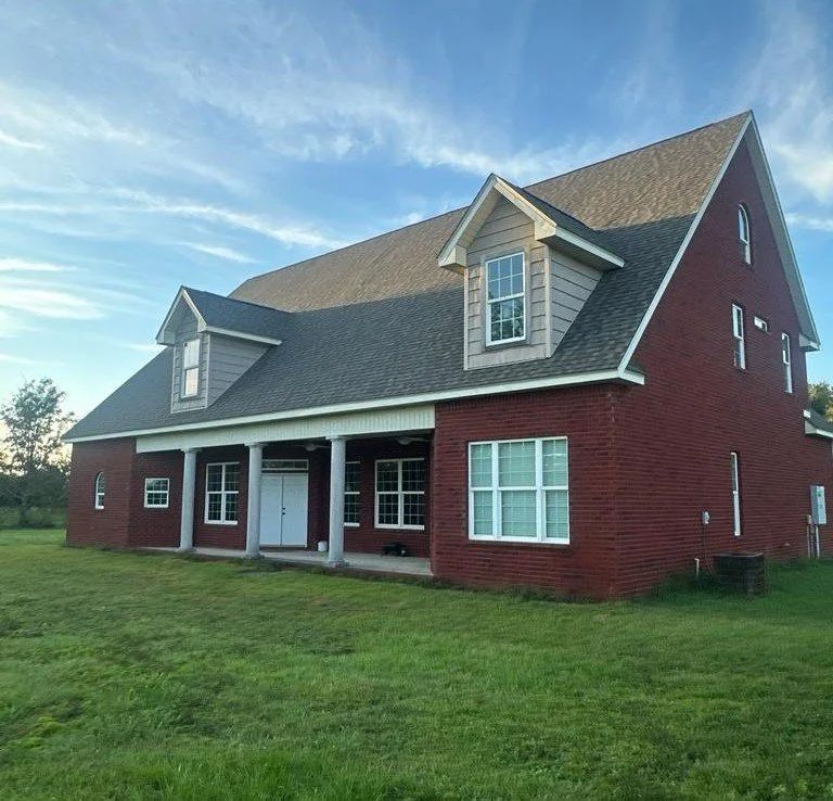 A large, two-story red brick house with a covered front porch, grey shingled roof, and two dormer windows.