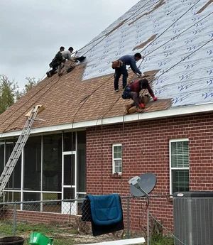 Four workers installing brown asphalt shingles on a steep residential roof with silver underlayment visible.