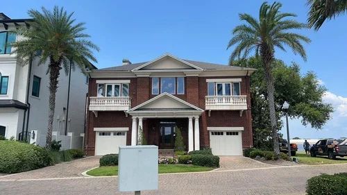 A two-story brick house with a white-pillared entrance, dual garages, and palm trees under a bright blue sky.