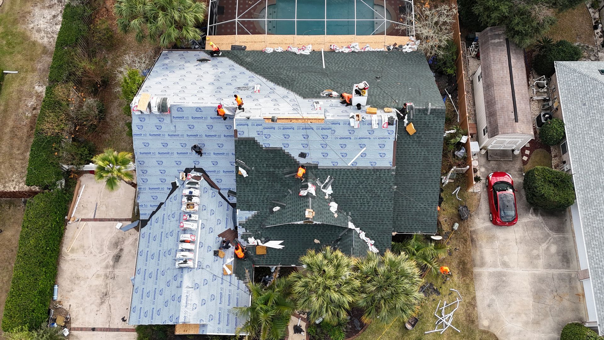 An aerial view shows workers on a residential roof installing shingles over gray underlayment in a suburban neighborhood.