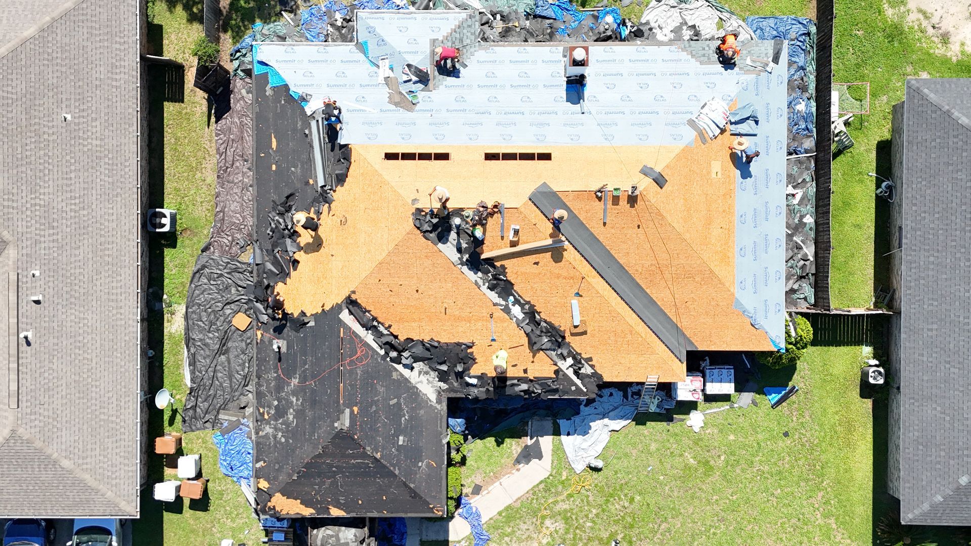 Aerial view of a house roof under repair, with exposed plywood, tar paper, and workers on top.
