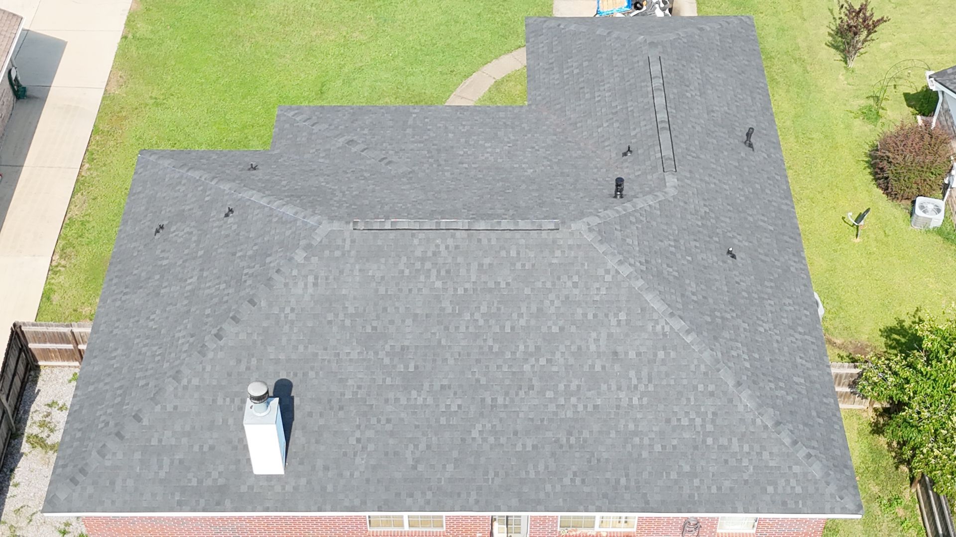 Aerial view of a gray shingled house roof with a chimney and surrounding green lawn.