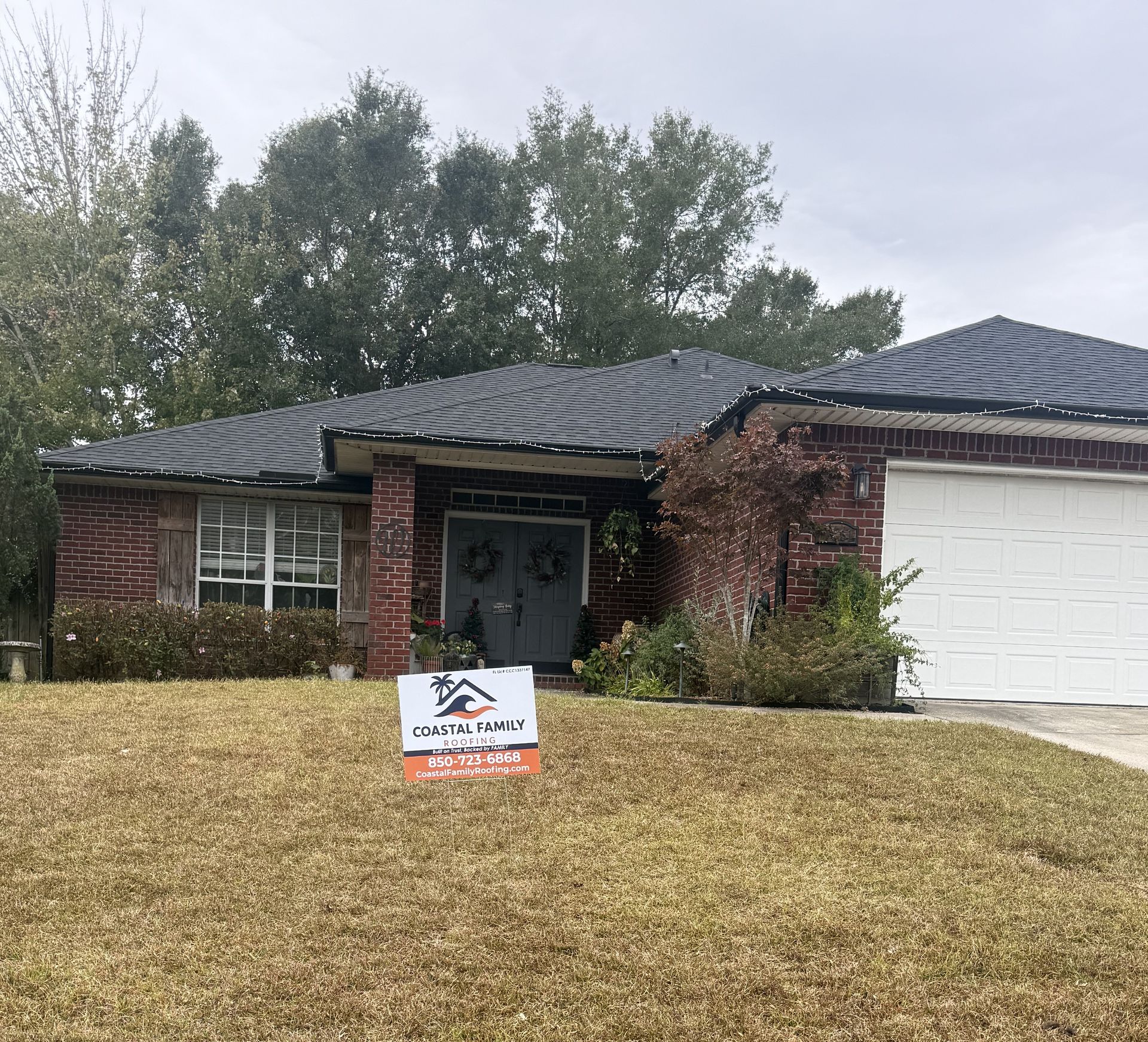 A brick, single-story house with a dark shingled roof, a white garage door, and a yard sign, set against a tree-lined sky.
