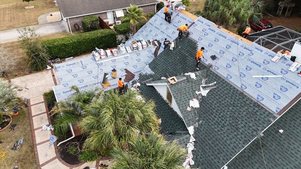 Workers installing blue roofing underlayment on a house roof, viewed from above.