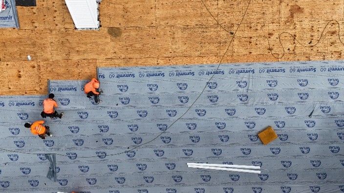 Three workers in orange gear install roofing underlayment on a wooden roof. Three workers in orange gear install roofing underlayment on a wooden roof.