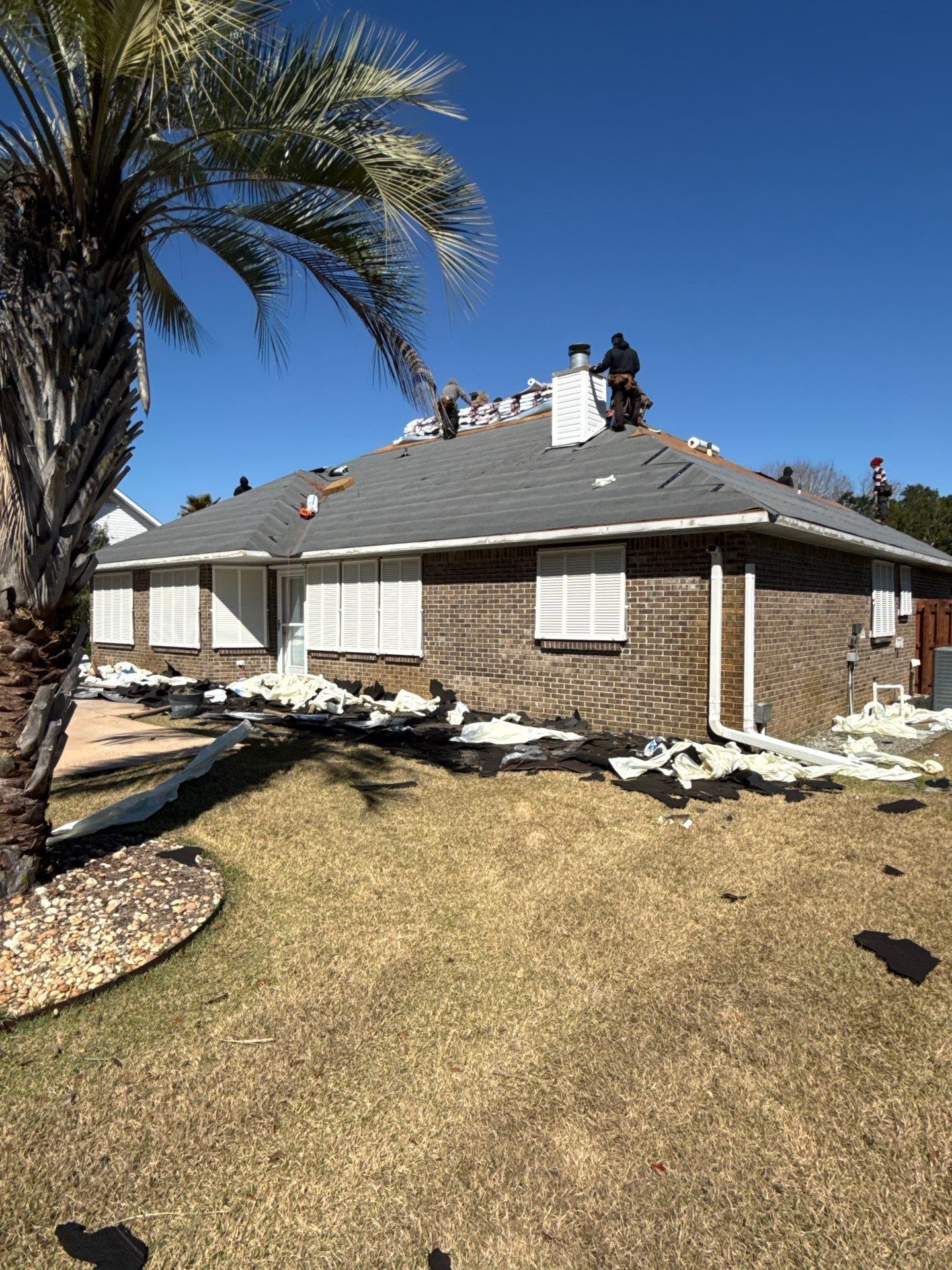 Overhead view of a house roof with dark shingles. Sections covered with tarps; trees below.