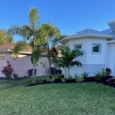 House exterior with palm trees, green grass, and flower beds under a blue sky.