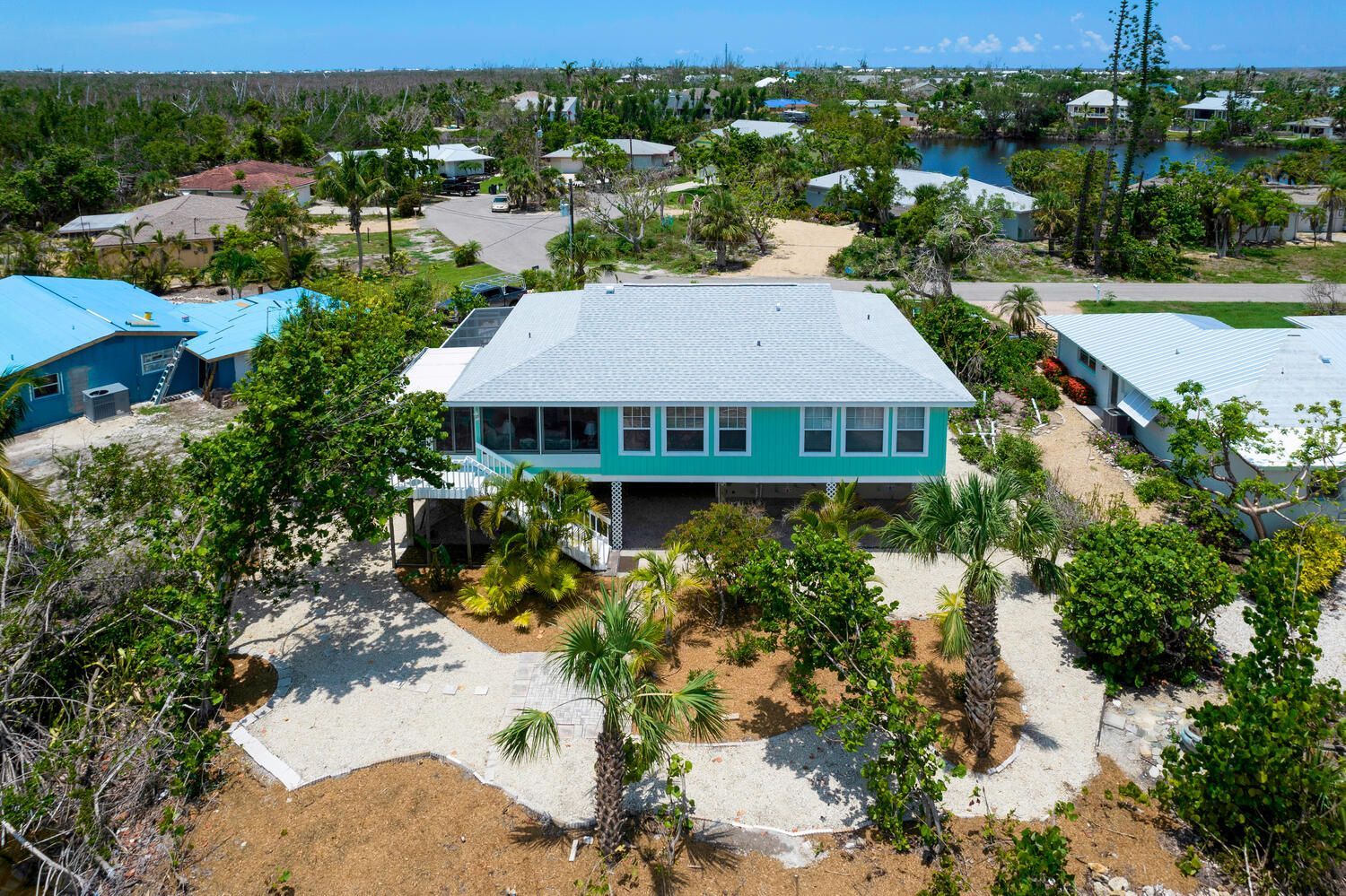Aerial view of a teal beach house with a white roof surrounded by greenery, near other homes.