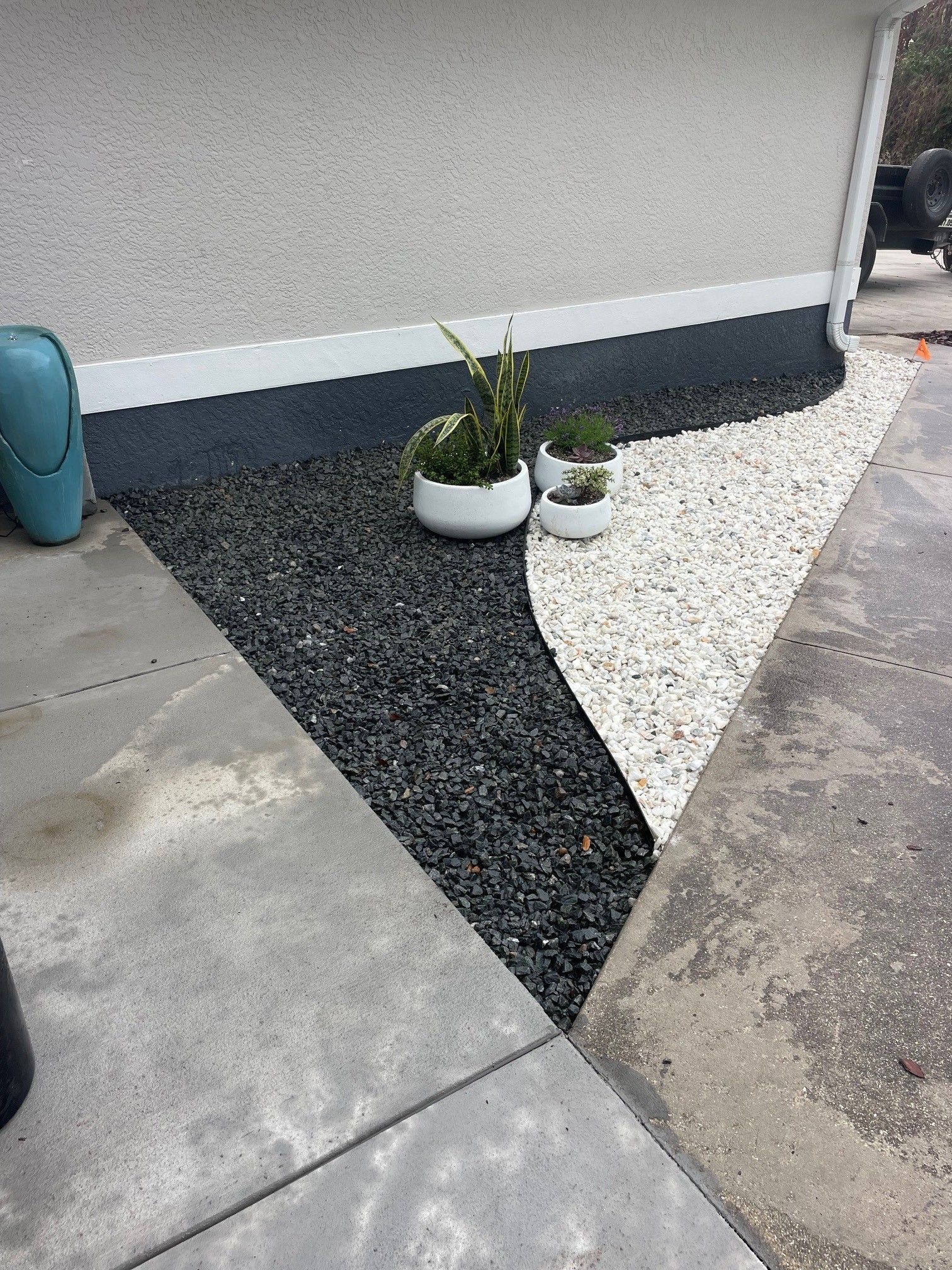 A triangular landscaping area features a curved split of black and white gravel, with three potted plants placed atop.
