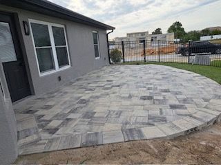 Gray paver patio adjacent to a gray house with a black door and windows. Black fence in the background.