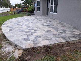 Gray paver patio extending from a gray house; grassy lawn, fence, and trees in background.
