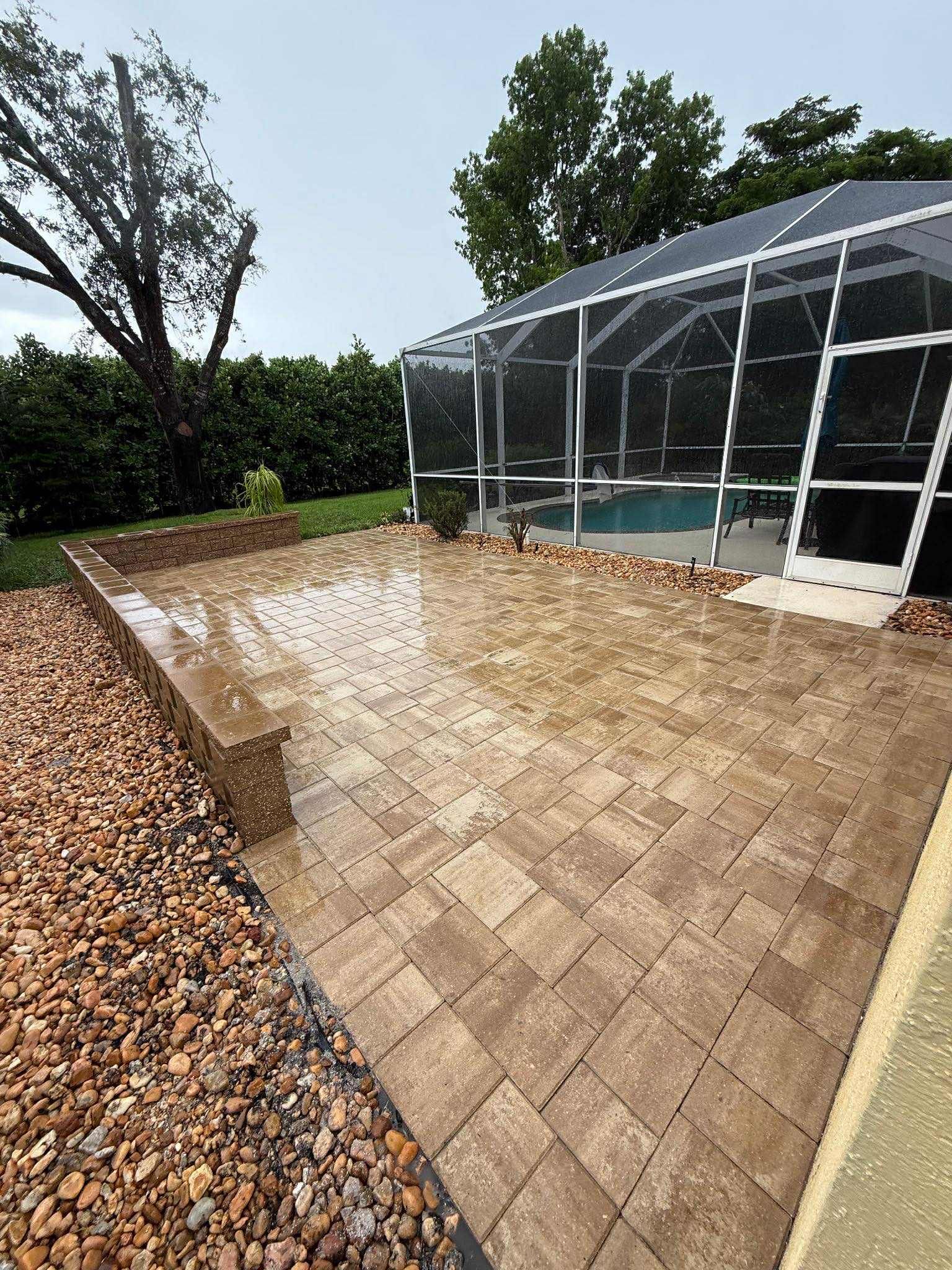 Paver patio with steps, pool enclosure, gravel border, and surrounding greenery. Overcast day.