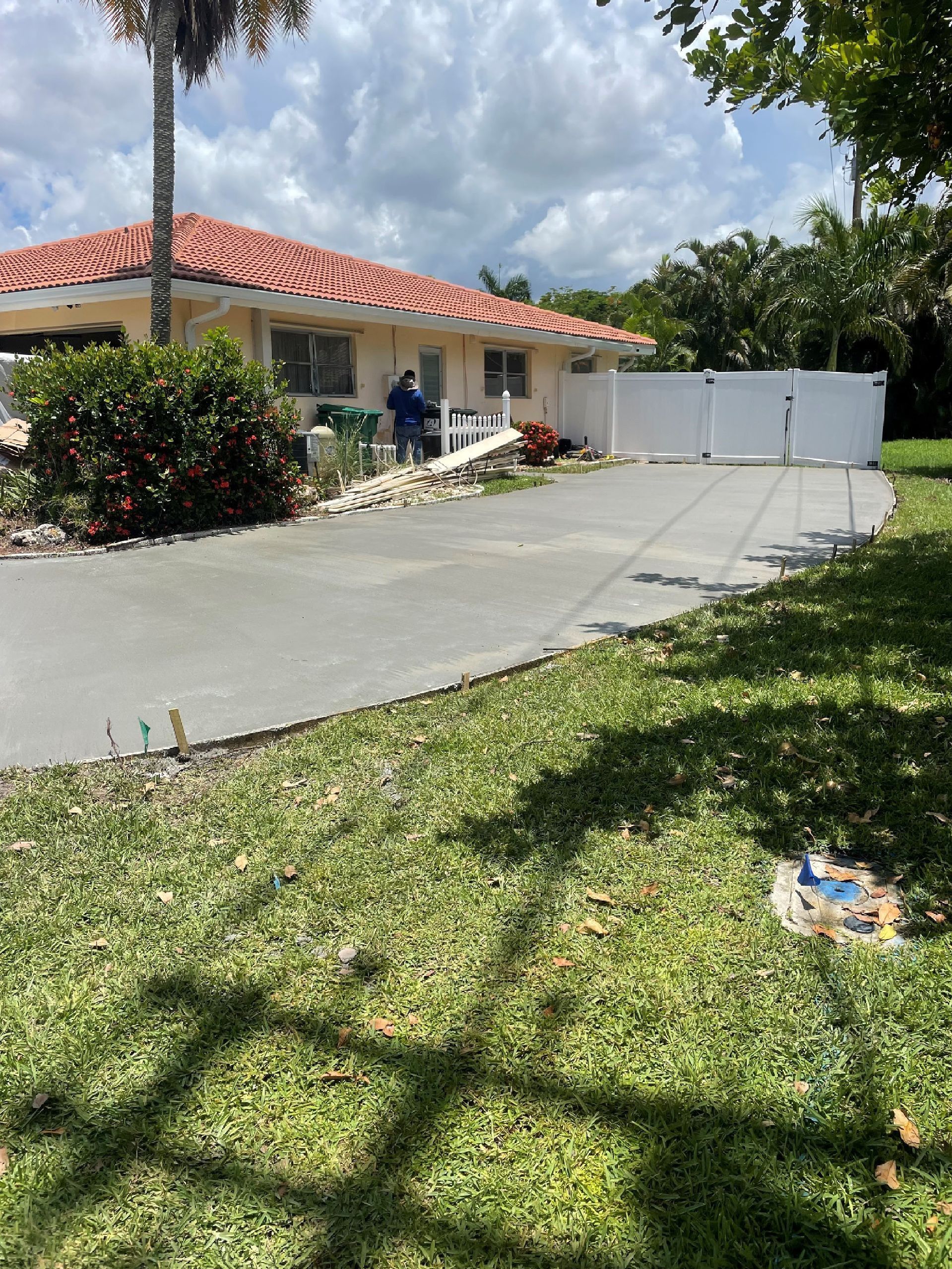 A person stands near a home with a driveway, surrounded by a white fence and green lawn.