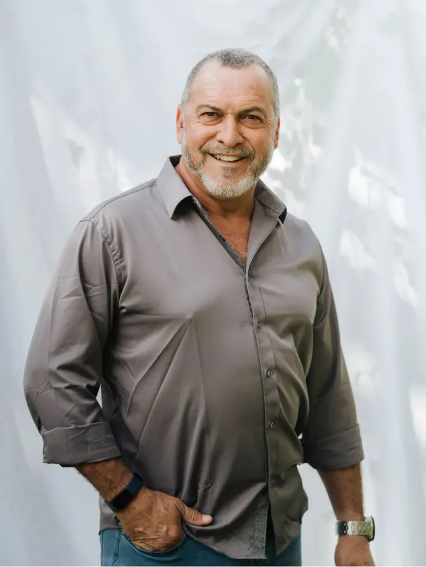 Man with grey beard and shirt smiles, standing outside.