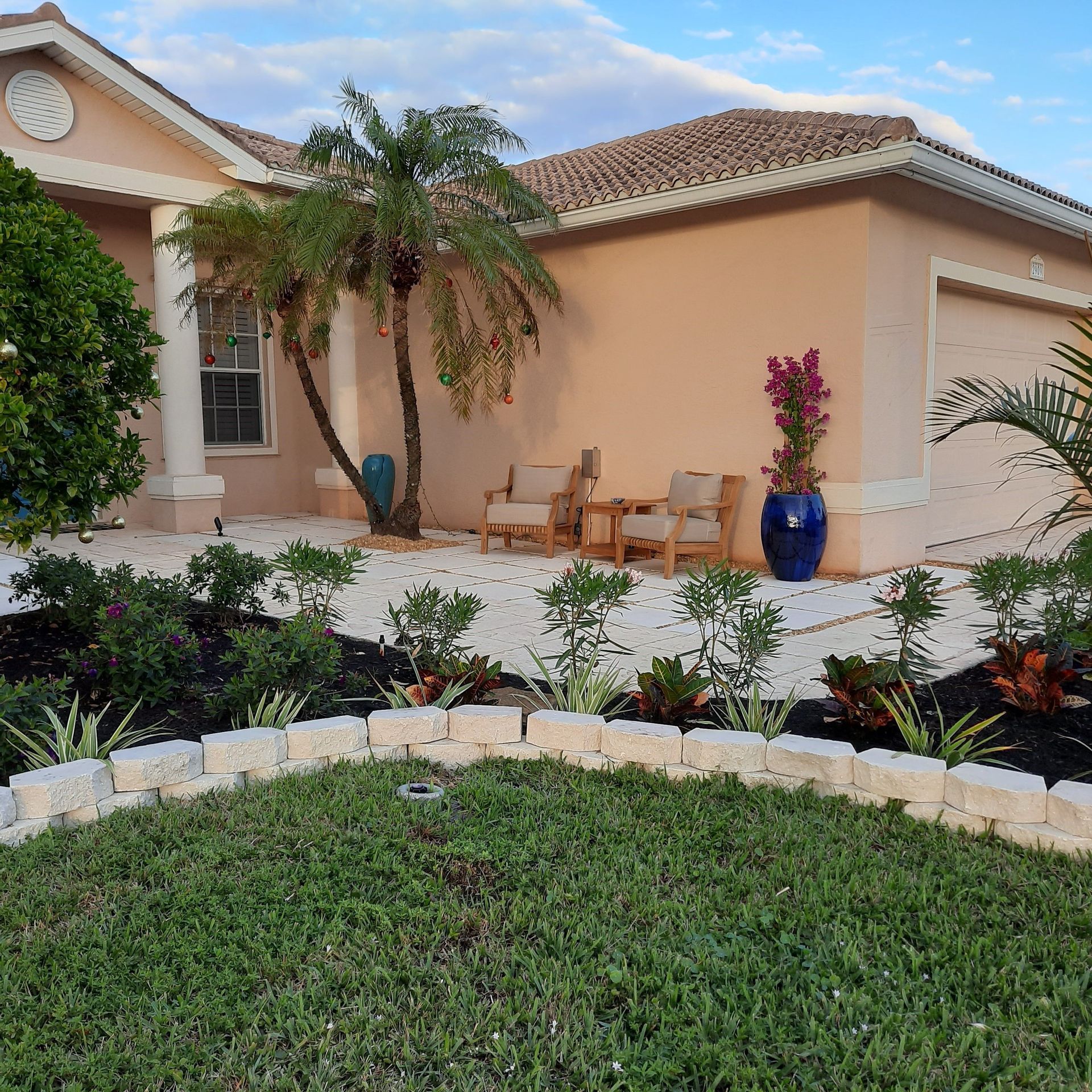 Landscaped front yard with palm tree, potted flowers, and tan stucco house with tiled roof