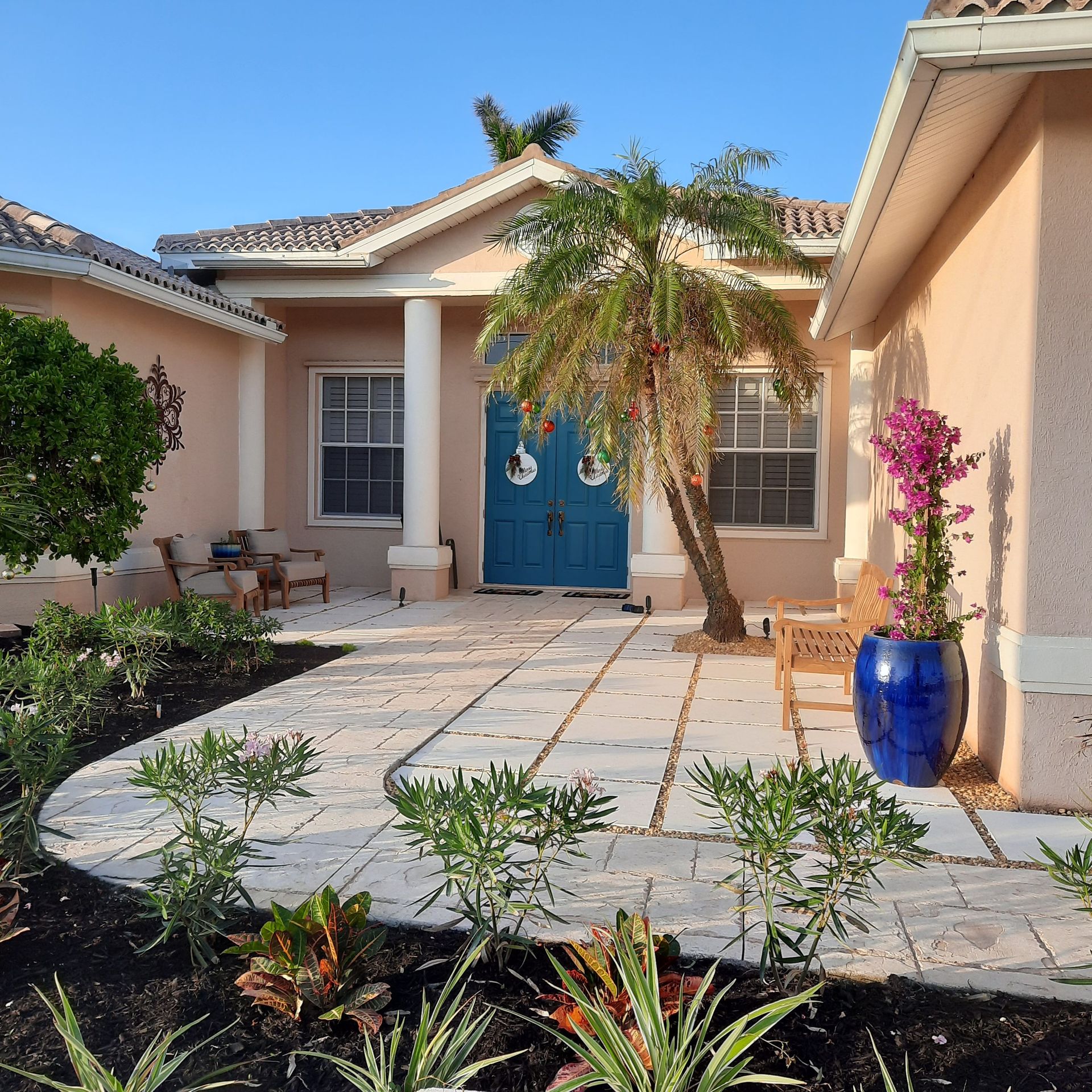 Front courtyard of a beige house with blue door, potted flowers, and a tiled walkway under a small tree