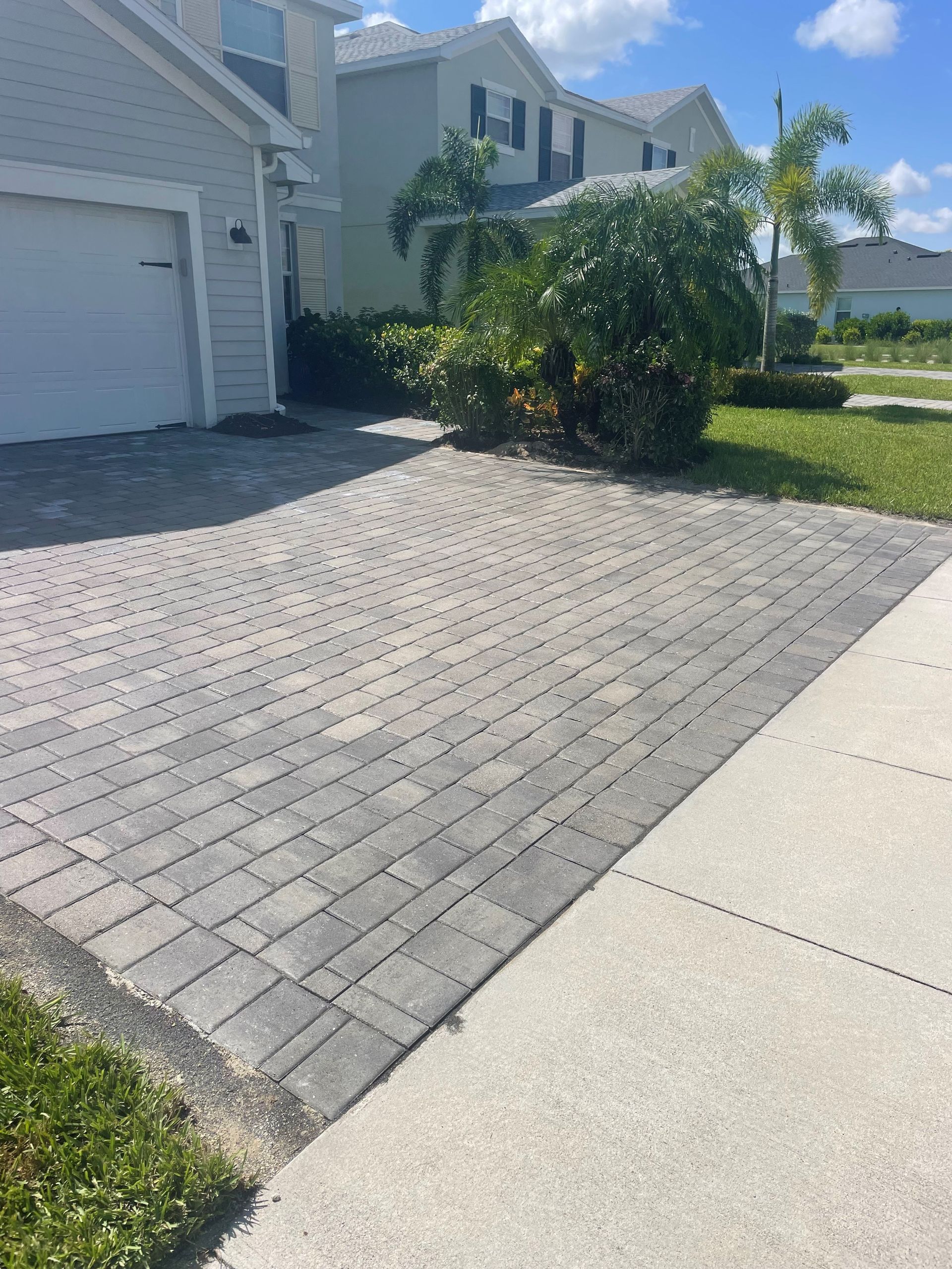 Brick driveway leading to a house, sidewalk on the right, green bushes and grass.