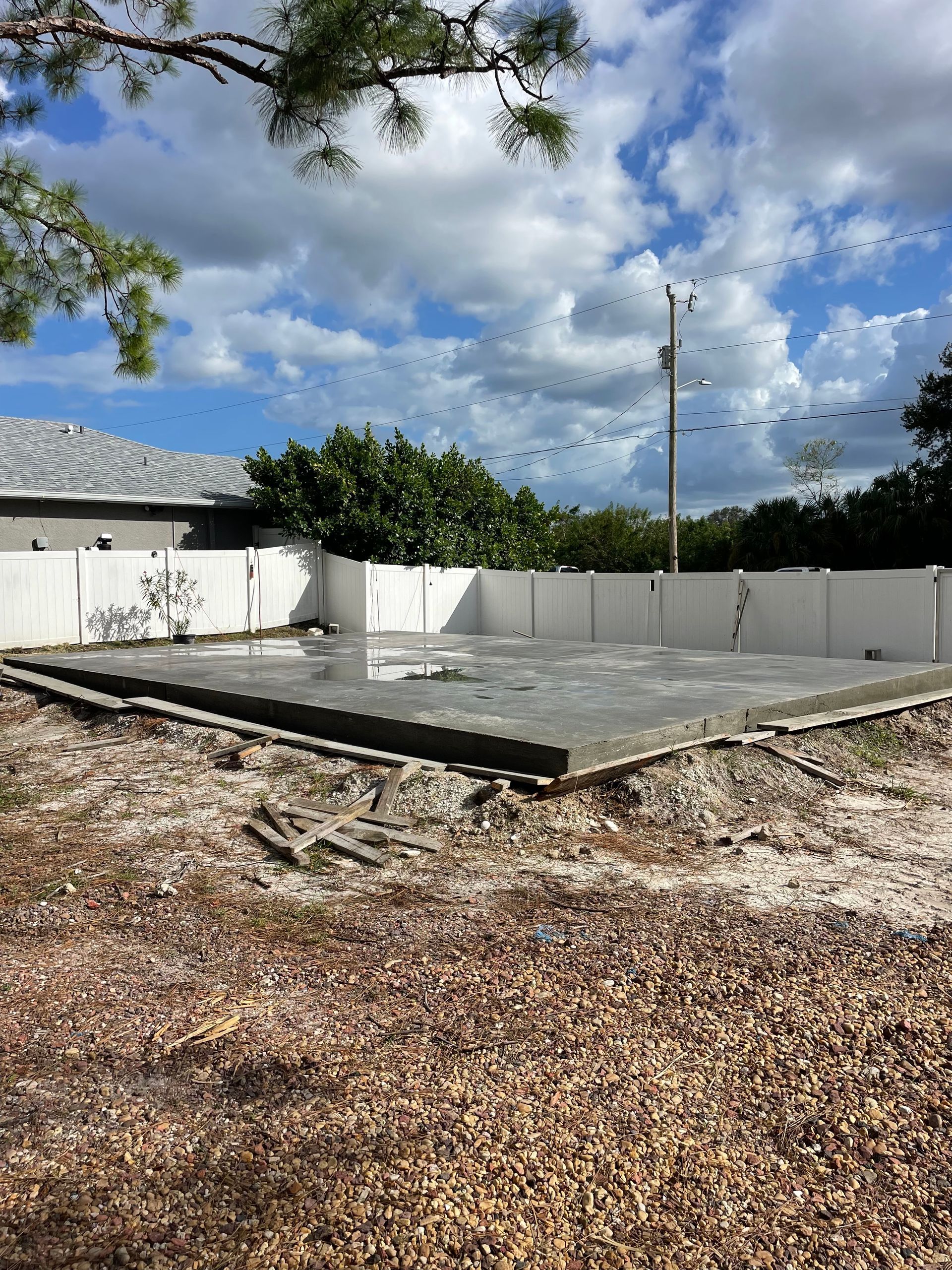Concrete foundation in backyard, surrounded by white fence, overcast sky overhead.