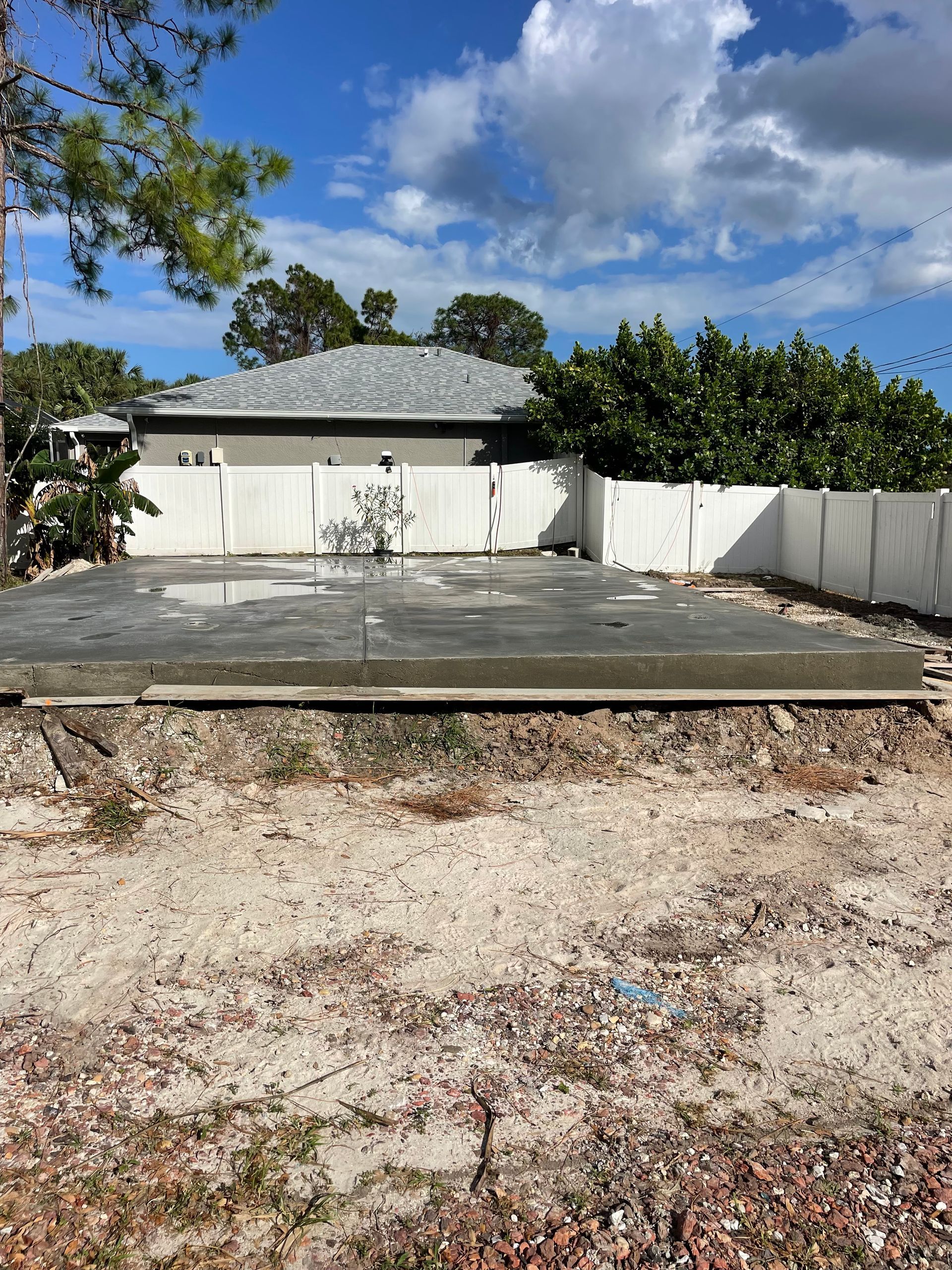 Concrete slab and gravel yard in front of a house with a white fence and blue sky.