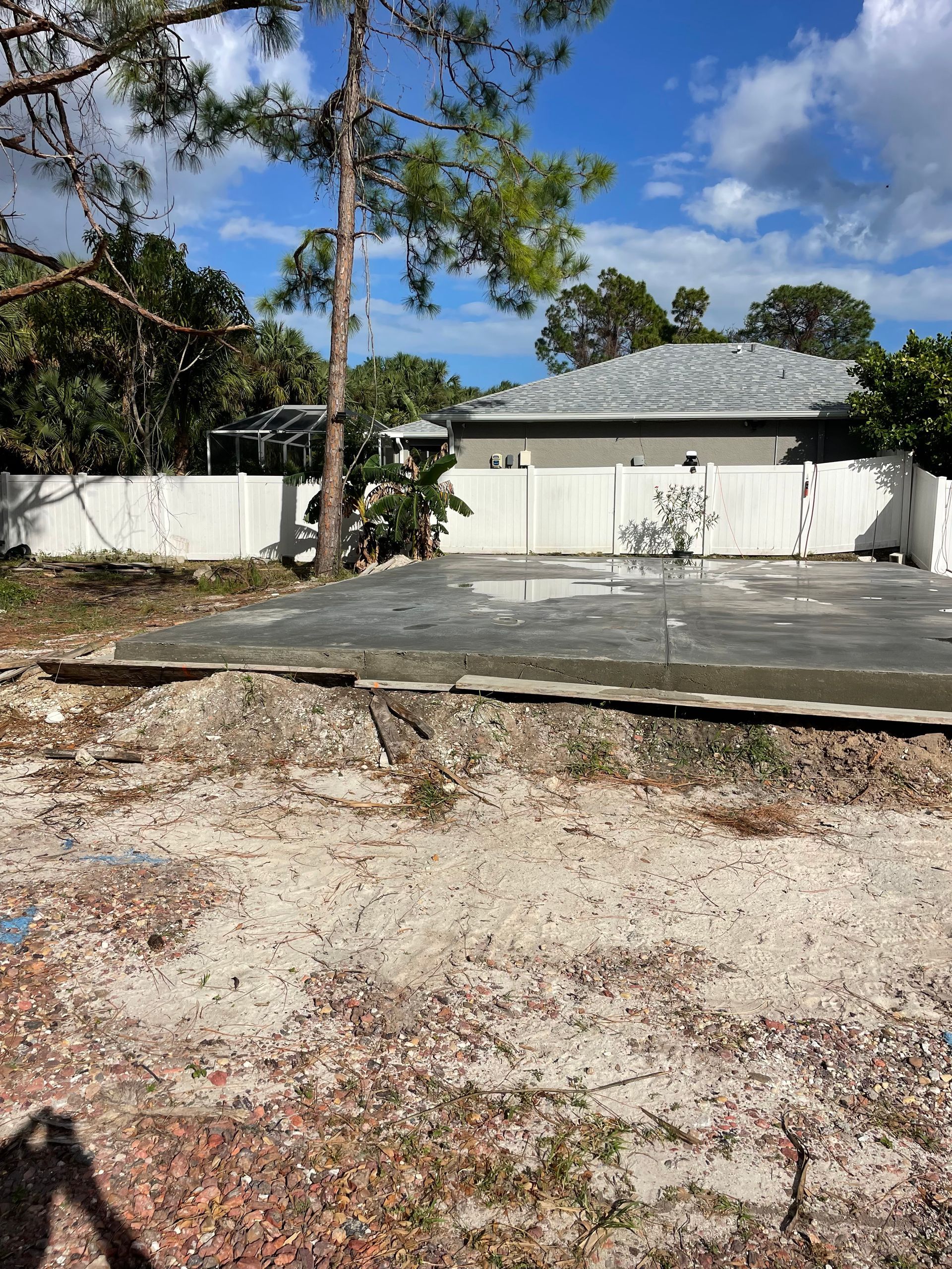 A partially constructed site, possibly a pool. White fence behind, trees and a house in the background on a sunny day.