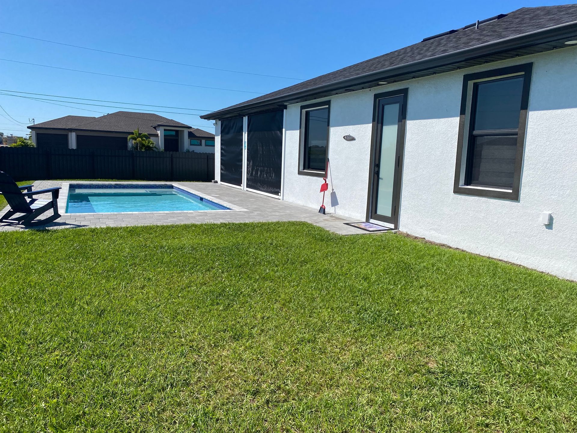 Backyard with a pool, green lawn, and white house with black trim under a clear blue sky.