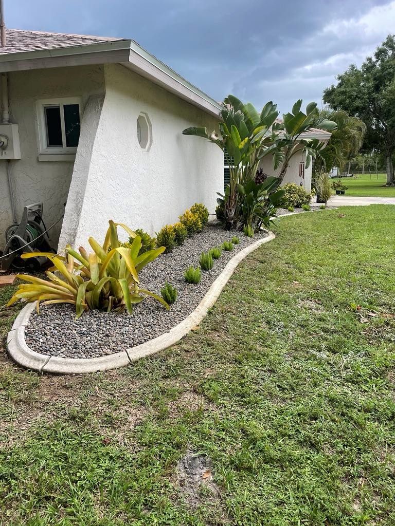 A house with a curving flower bed filled with plants and dark pebbles, framed by a concrete border.