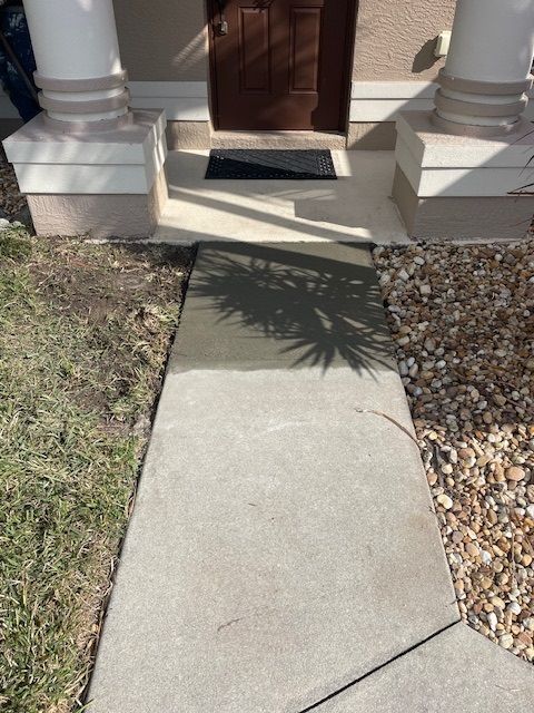 Concrete walkway leading to a doorway with a welcome mat, flanked by grass and gravel.