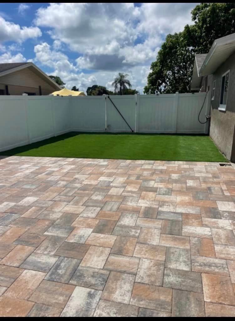 Backyard patio with brick pavers, green lawn, white fence, and cloudy sky