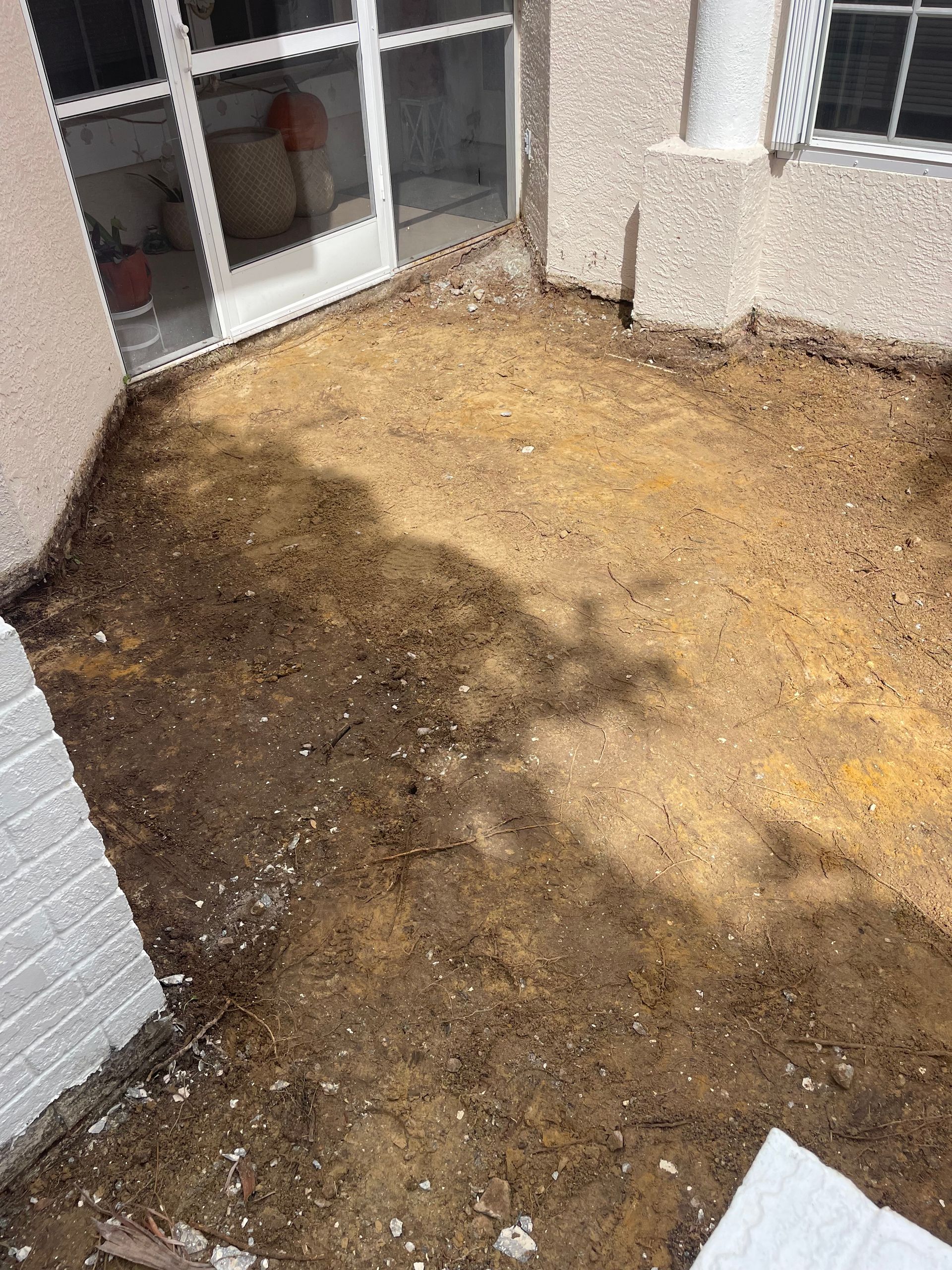 Dirt patch between a house and a screened porch, with shadows and light-colored brickwork on the side.